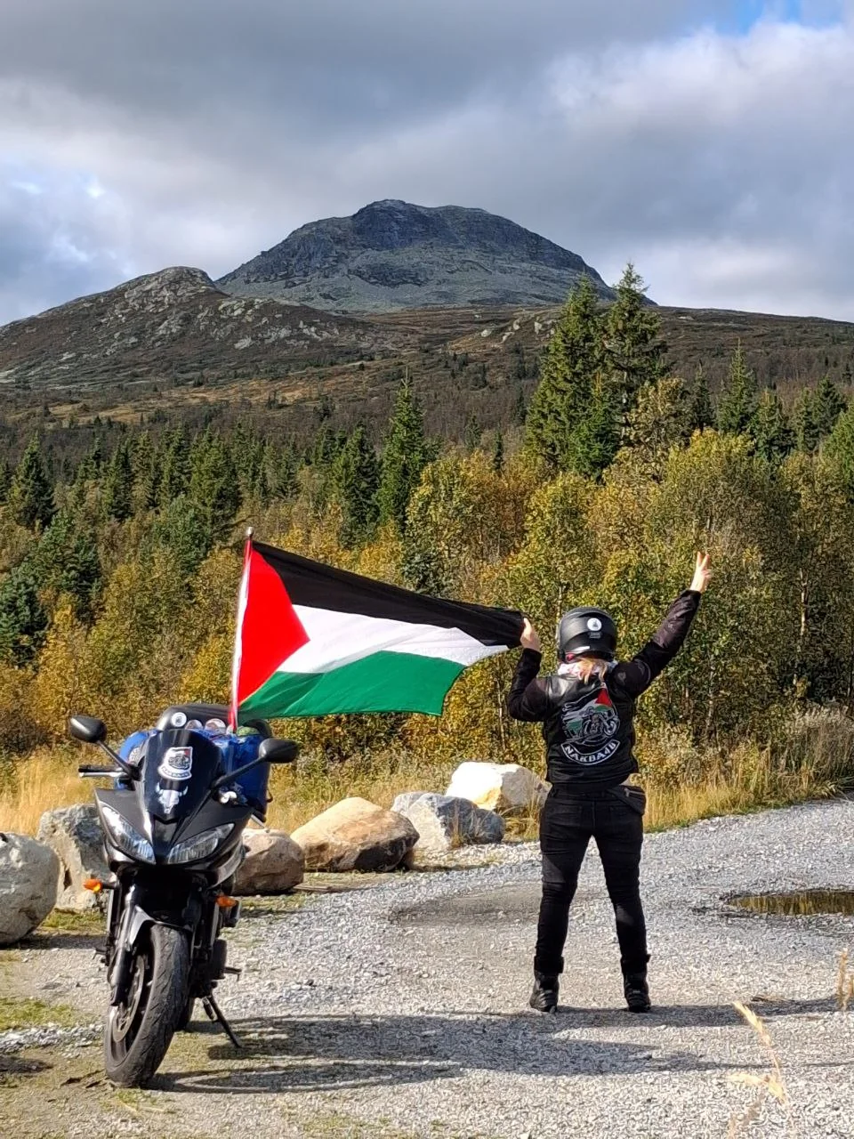 Motorist s palestinsko zastavo ob motorju v gorski pokrajini.